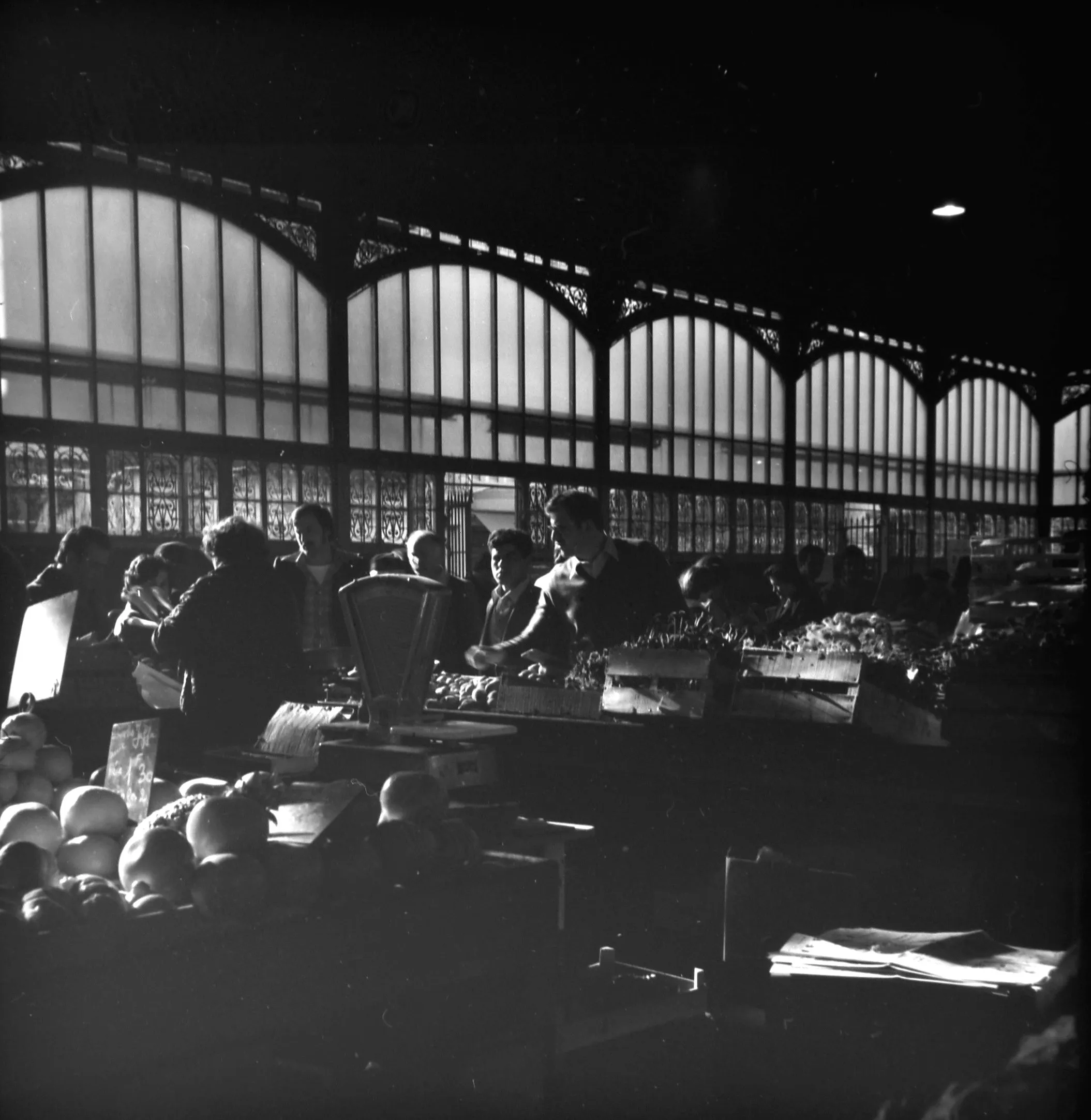 Black and white photo of a busy indoor market with people shopping and vendors behind wooden crates of produce, illuminated by large arched windows.