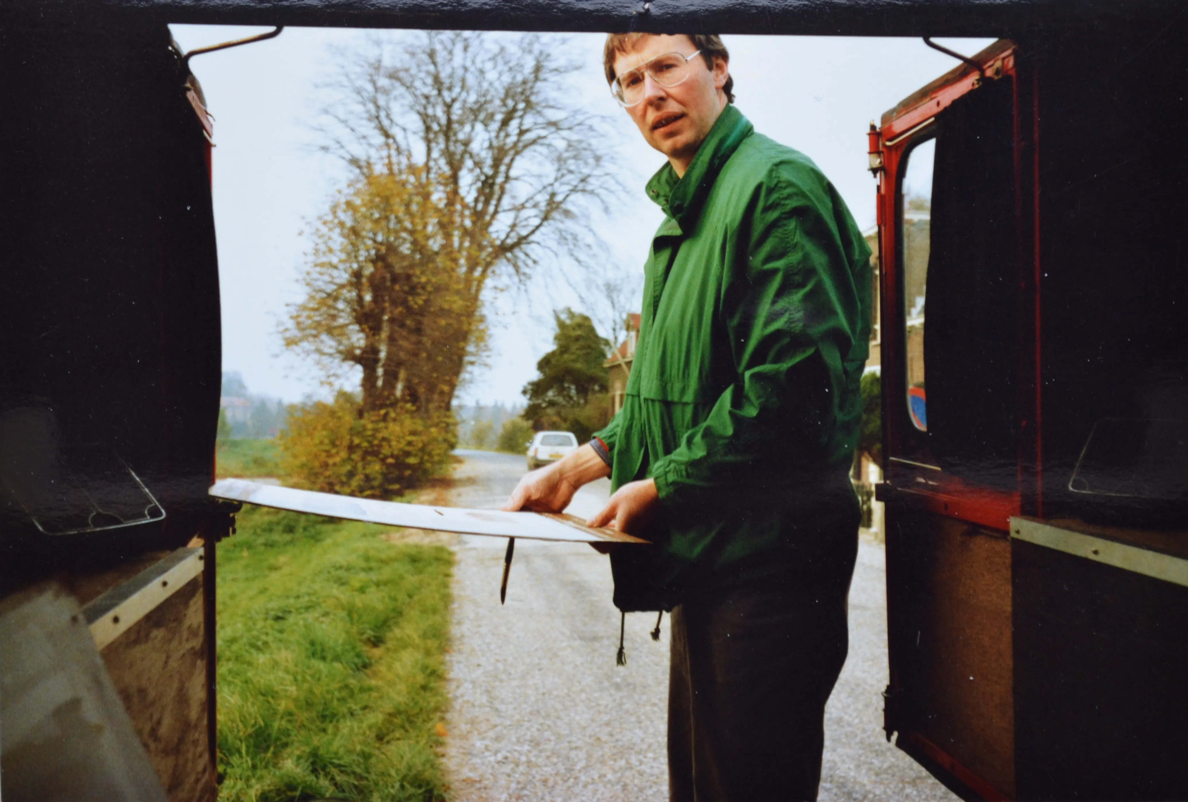 Man in green jacket standing outside a vehicle, holding a painting board with a brush attached, on a country road with trees and a car in the background.