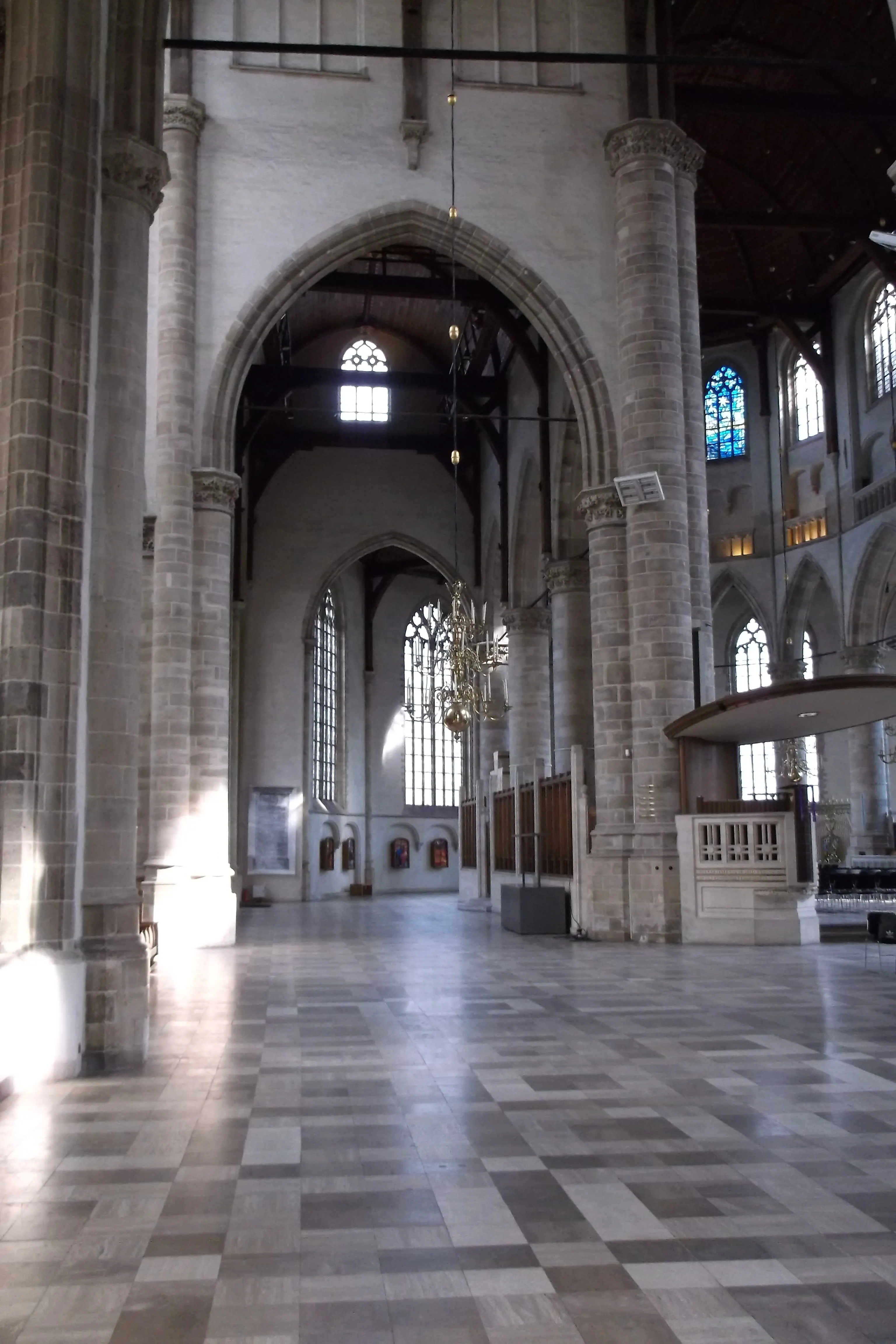 Interior of a large church with tall stone columns, arched windows, chandeliers, and a checkered tile floor.