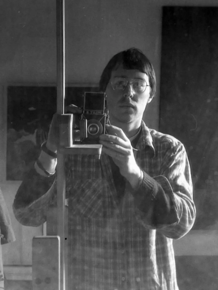 Black and white photo of a man wearing glasses and a checkered shirt, taking a mirror selfie with a vintage camera.