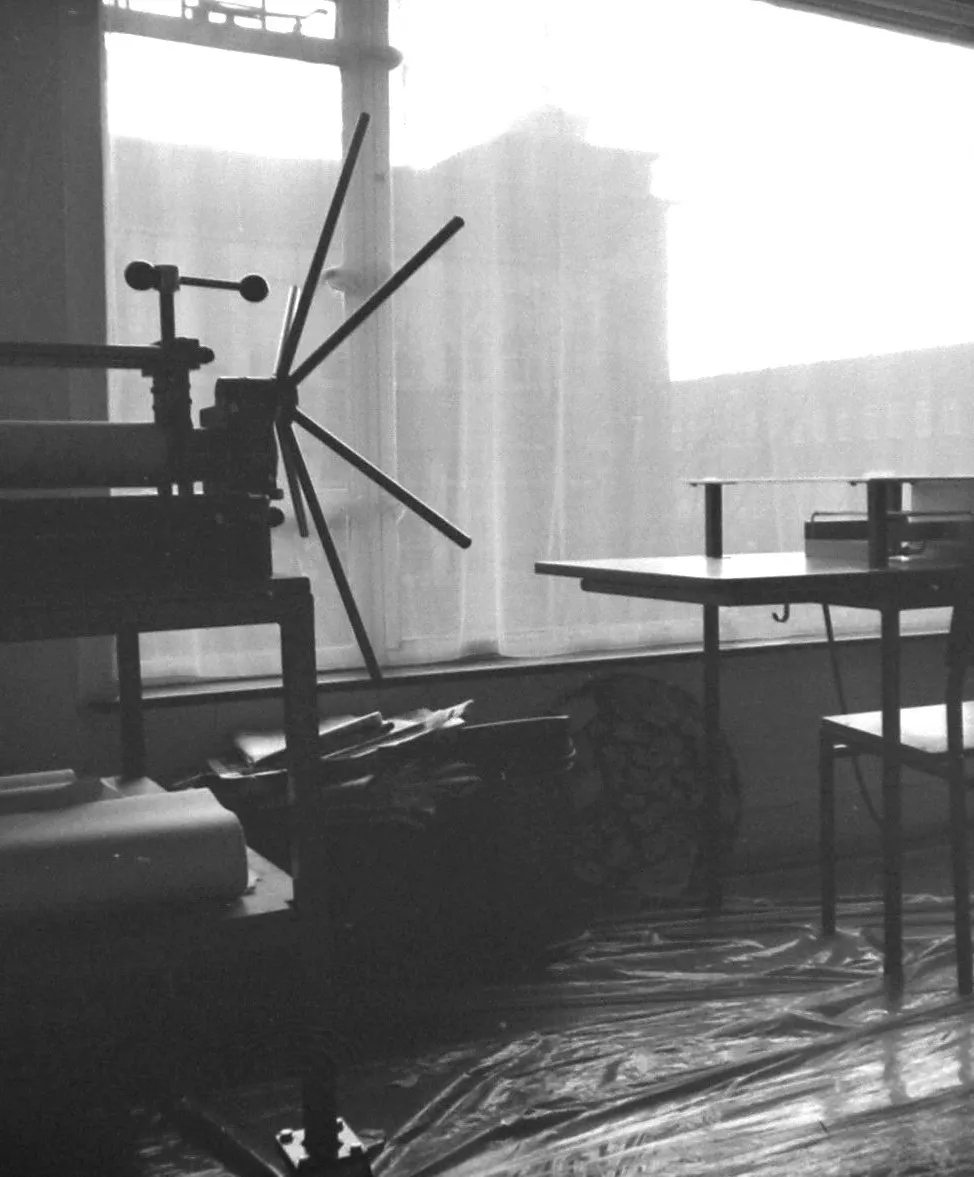 Black-and-white photo of an industrial printing press with a large spoked wheel next to a window, a table, and a chair in a workspace.
