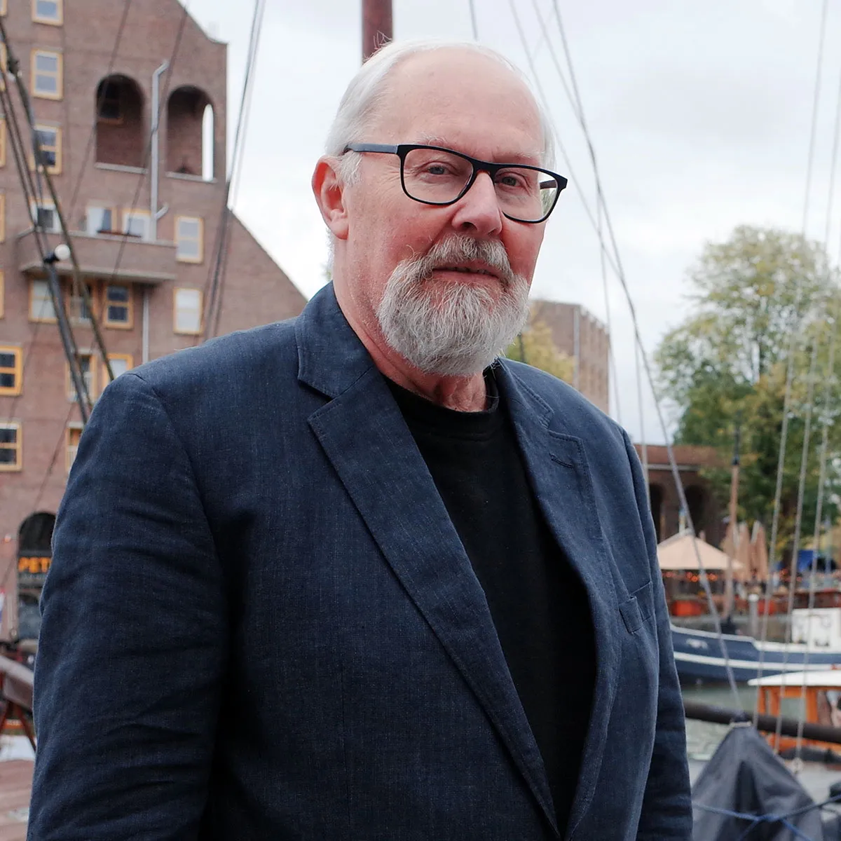 Older man with white hair, beard, and glasses wearing a dark blazer and black shirt standing near boats in a harbor.