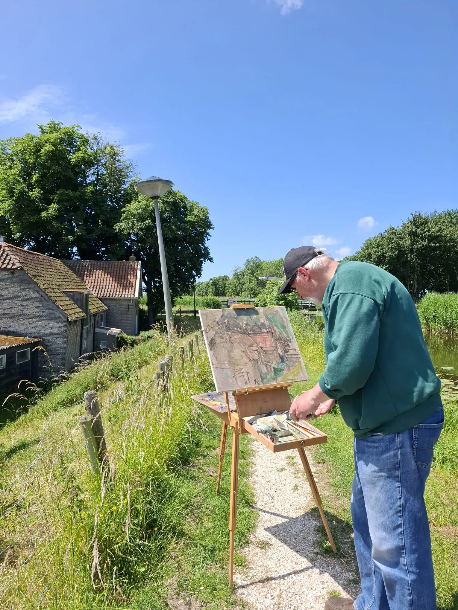 Elderly man wearing a green sweatshirt and cap painting outdoors on an easel beside a grassy path near houses and a pond under a clear blue sky.