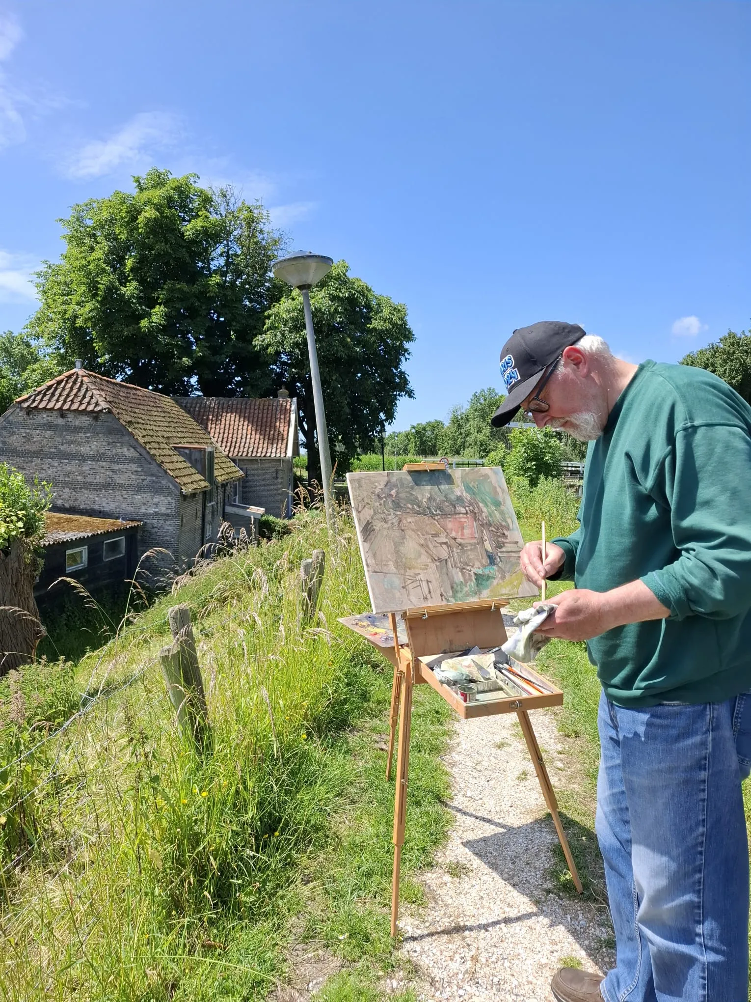 An elderly man painting outdoors on a wooden easel along a grassy path near rural houses under a clear blue sky.