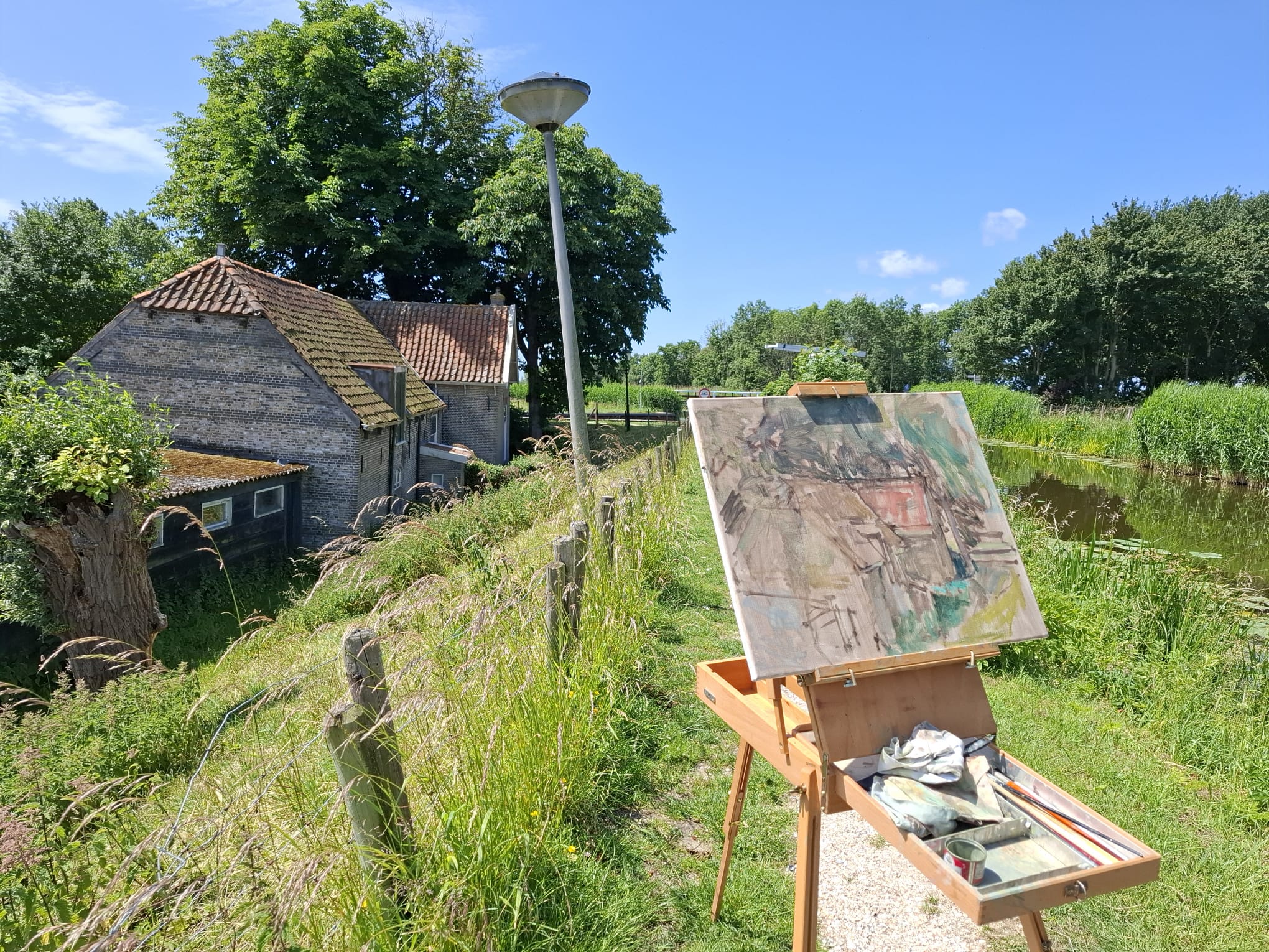 Outdoor easel with a landscape painting of a rustic house beside a pond under a clear blue sky.