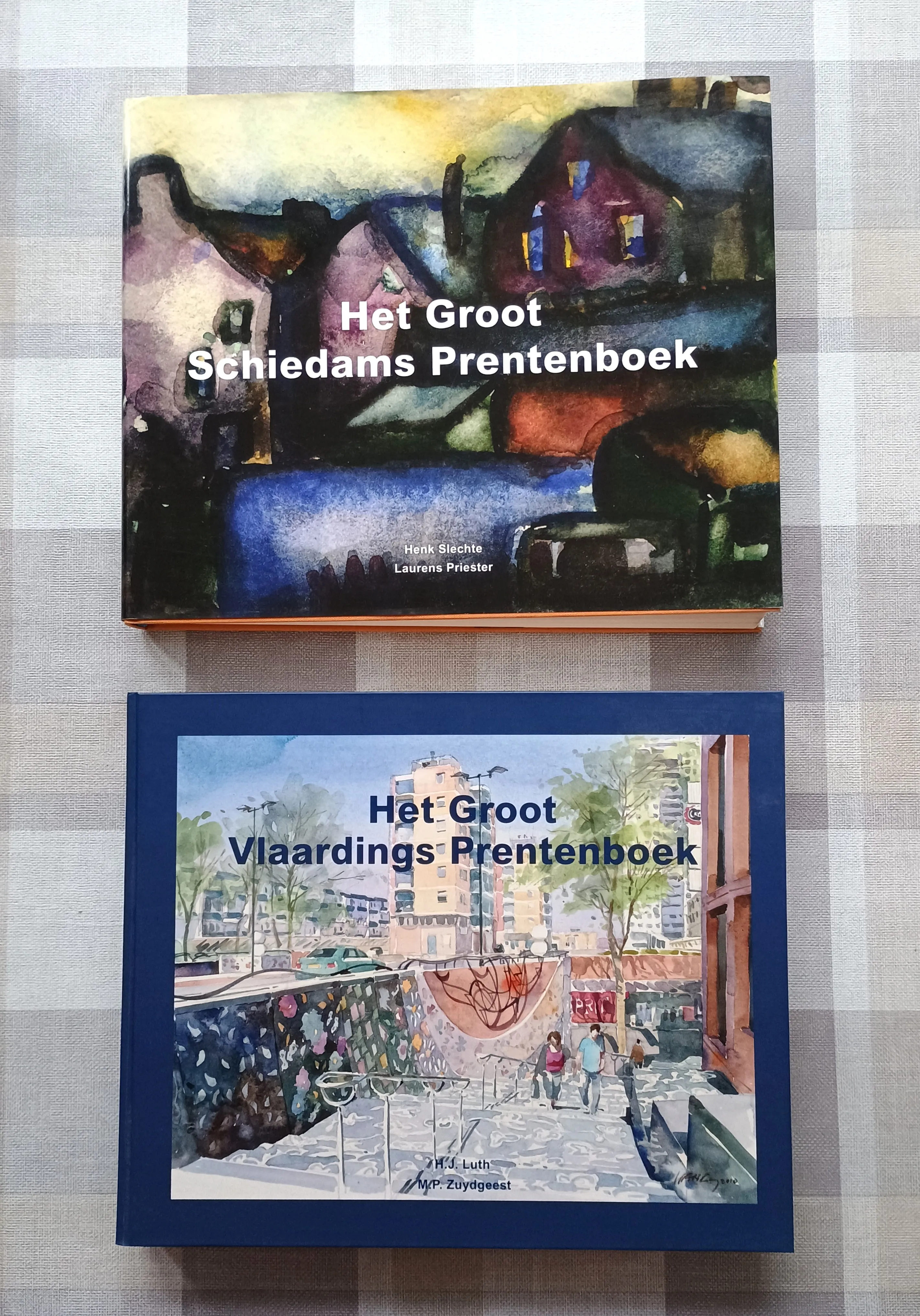 Two books on a checkered tablecloth: top book titled 'Het Groot Schiedams Prentenboek' with an abstract watercolor house scene; bottom book titled 'Het Groot Vlaardings Prentenboek' with a watercolor urban street scene featuring people and buildings.
