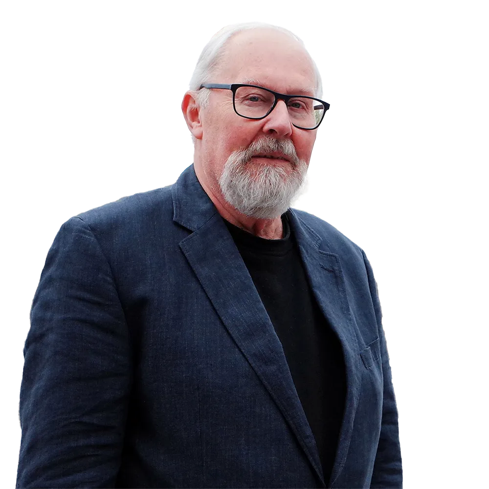 Older man with white hair and beard wearing black glasses, a navy blazer, and a black shirt, standing outdoors with buildings and boat masts in the background.