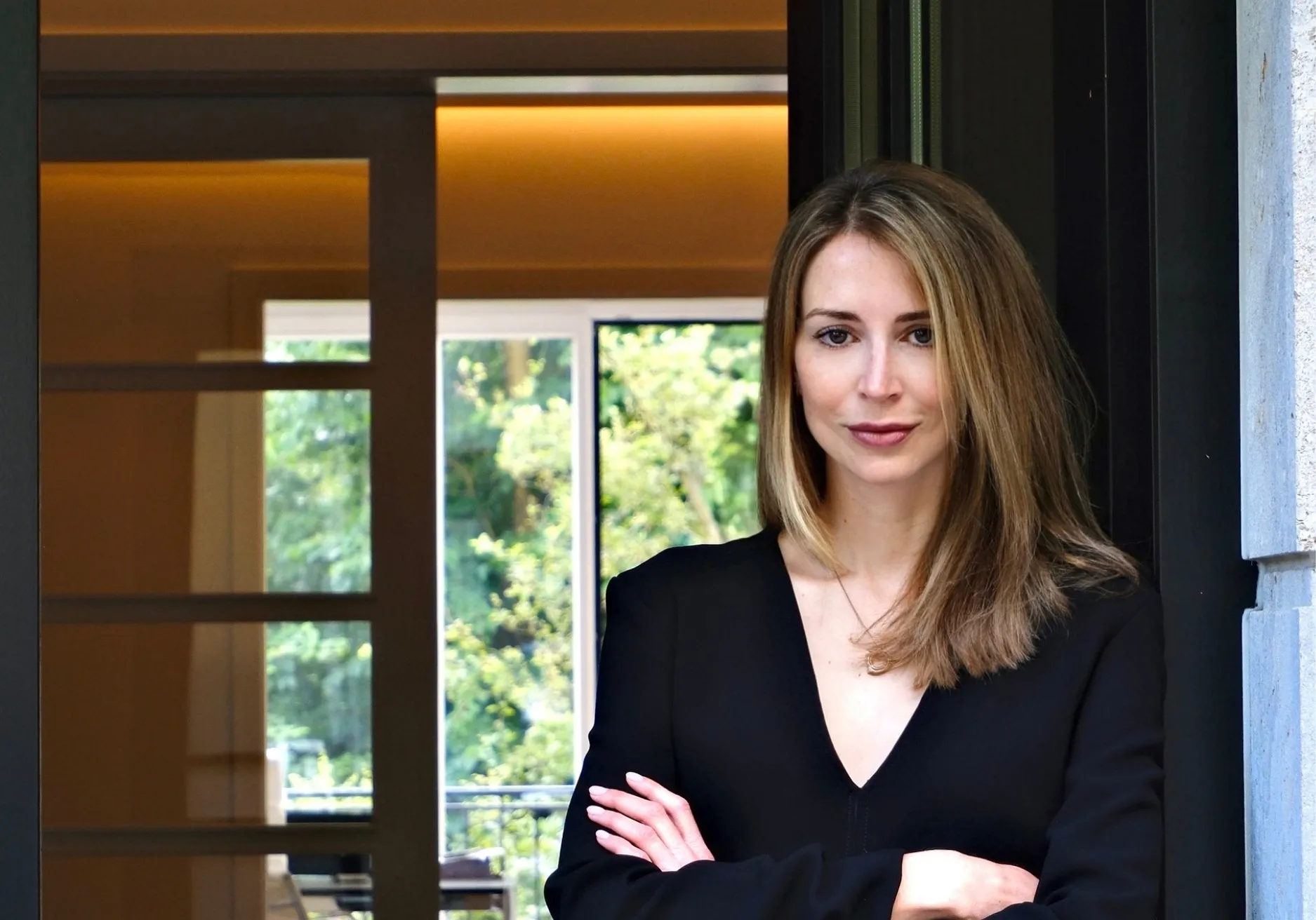 Dr. med. Lucija Grittern, geb Kröpfl – Fachärztin für Dermatologie - mit langen blonden Haaren und schwarzem Oberteil steht vor einem Fenster mit Blick auf grüne Bäume.