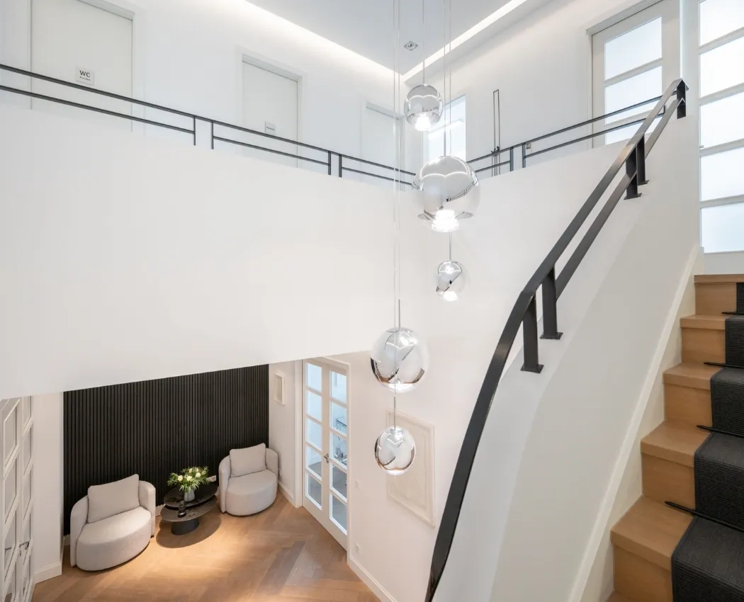 Moderner Innenraum mit Holztreppe, schwarzen Geländer, hängenden silbernen Kugelleuchten und zwei grauen Sesseln neben einem kleinen Tisch mit Blumen.