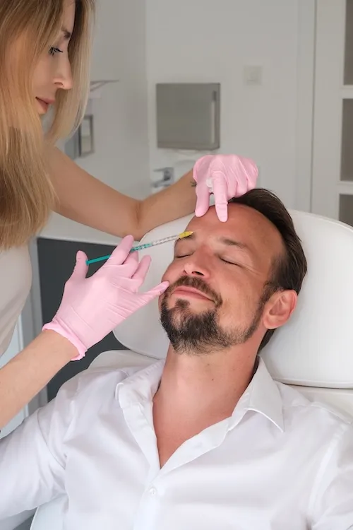 Dr. med. Lucija Grittern, geb Kröpfl – Fachärztin für Dermatologie - mit rosa Handschuhen injiziert einem entspannten Mann eine Behandlung in die Stirn.