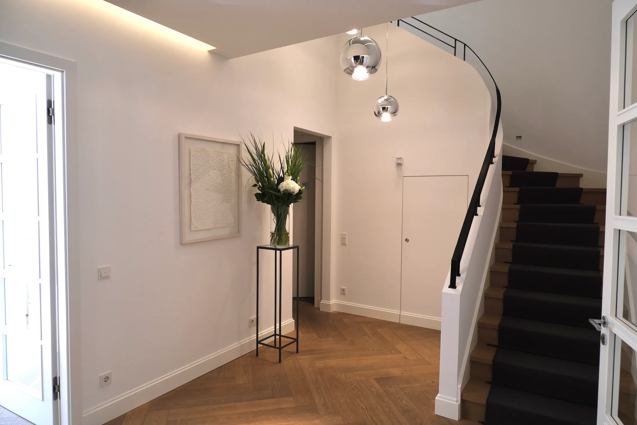 Moderner heller Flur mit Holzparkett, Treppe mit schwarzem Teppichläufer, großem Blumenarrangement auf einem schmalen schwarzen Gestell und zwei hängenden Kugelleuchten.