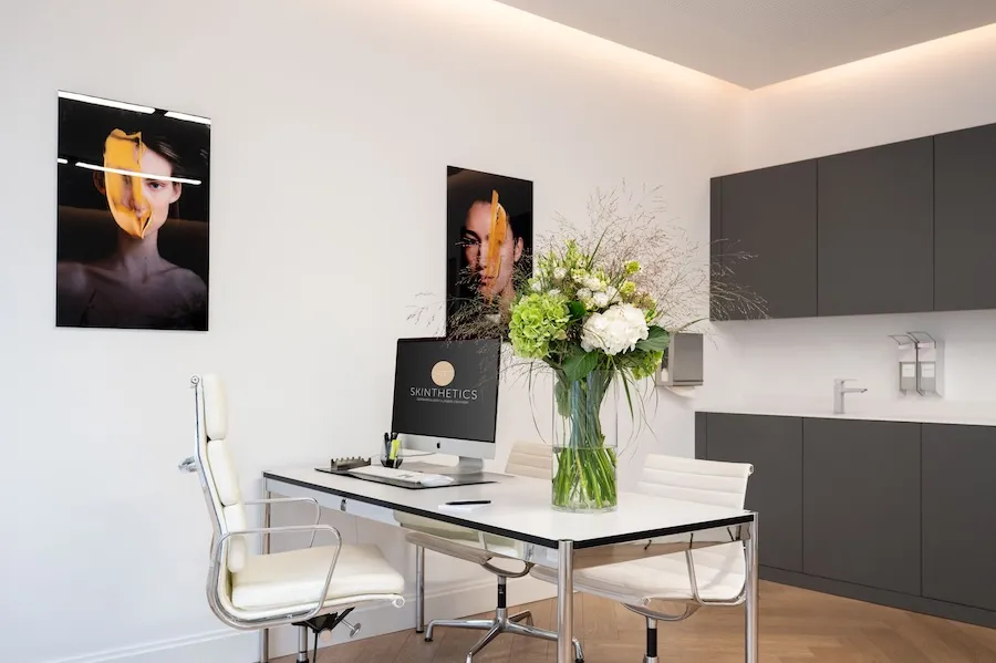 Moderner Behandlungsraum mit weißem Schreibtisch, drei weißen Stühlen, einem großen Blumenstrauß und einem Computerbildschirm mit der Aufschrift 'SkinThetics'.