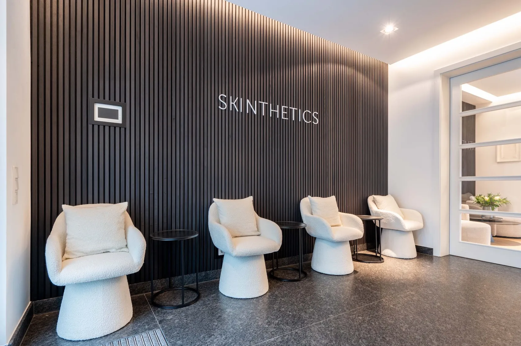 Wartebereich mit vier modernen, weißen gepolsterten Sesseln vor einer schwarzen Wand mit vertikalen Linien und Schriftzug 'SKINTHETICS'.