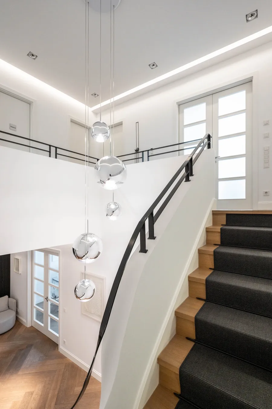 Moderner Treppenaufgang mit Holzstufen und schwarzem Teppichläufer, weiße Wände und silberne Kugel-Pendelleuchten.