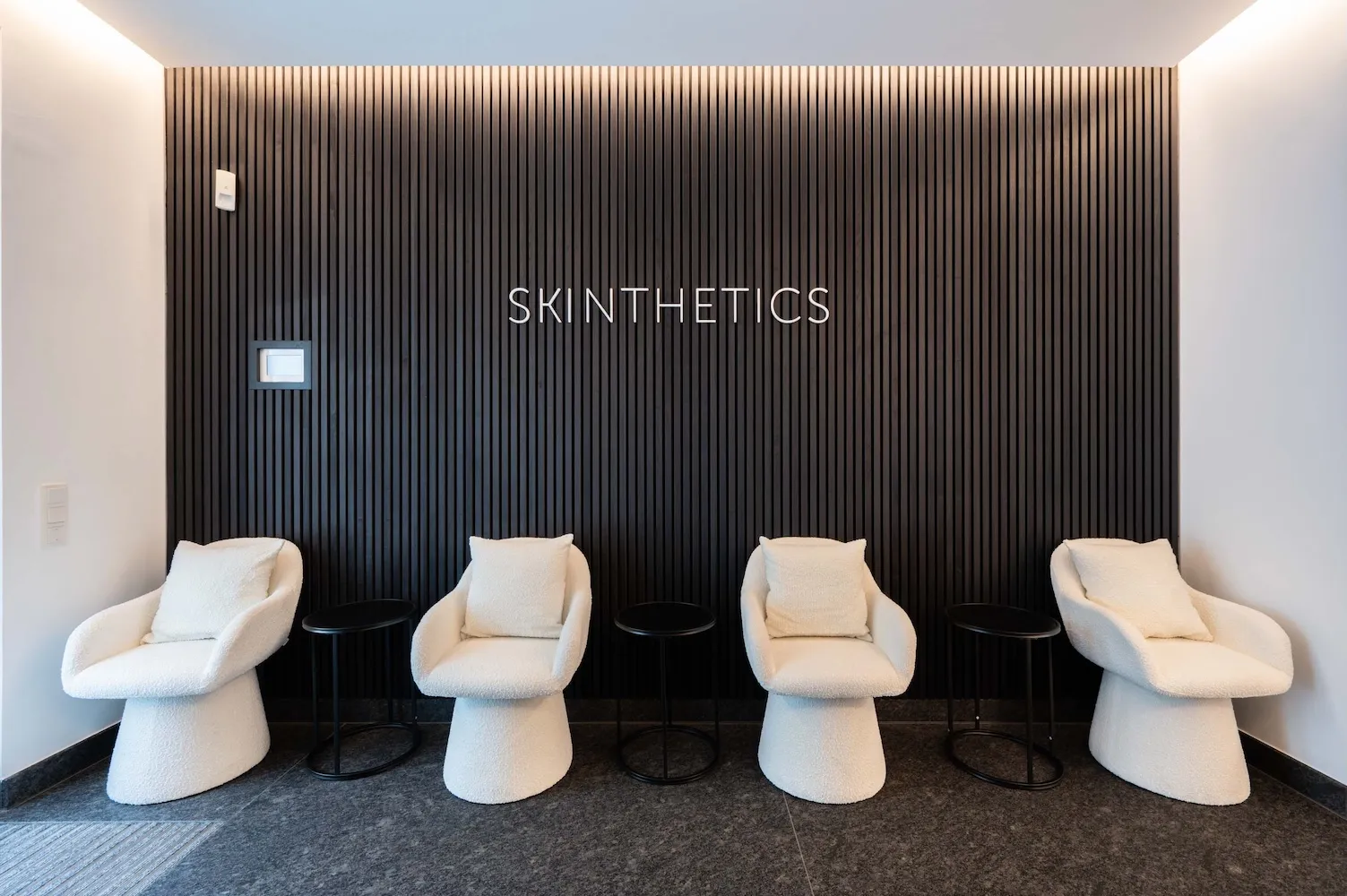 Vier weiße gemütliche Stühle mit Kissen vor einer schwarzen, vertikal geriffelten Wand mit dem Schriftzug 'SKINTHETICS'.