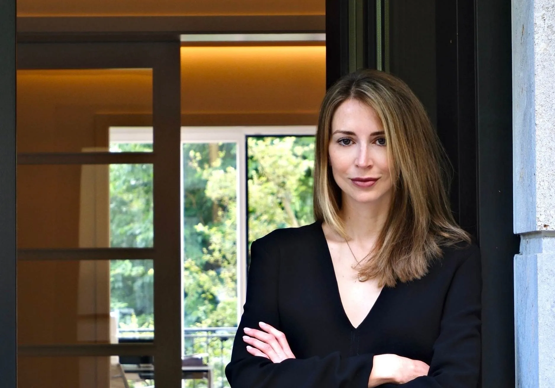 Dr. med. Lucija Grittern, geb Kröpfl – Fachärztin für Dermatologie mit langen blonden Haaren trägt ein schwarzes Oberteil und steht mit verschränkten Armen vor einem Fenster mit Blick auf grüne Bäume.