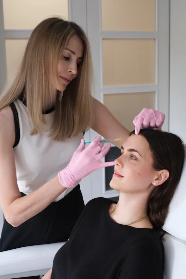 Ärztin mit rosa Handschuhen injiziert einer entspannten Patientin eine Spritze in die Stirn.