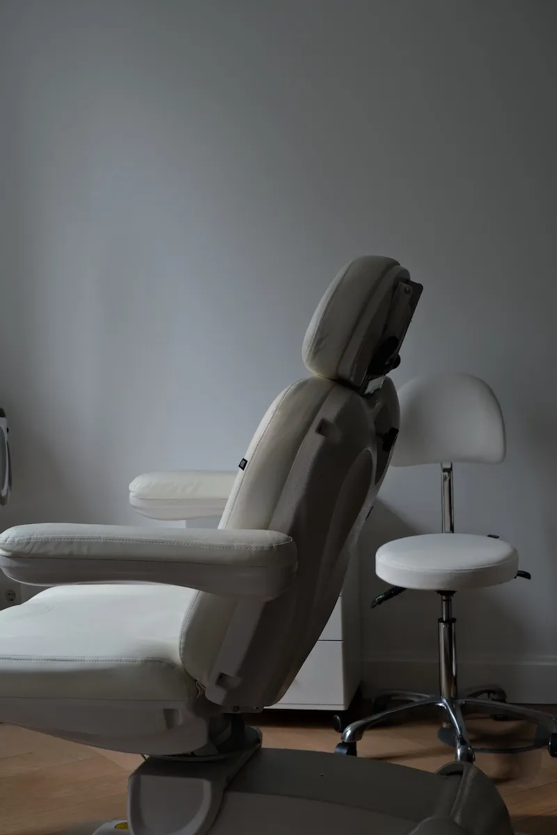 Ein weißer, verstellbarer Arzt- oder Behandlungsstuhl neben einem weißen Drehstuhl vor einer grauen Wand.