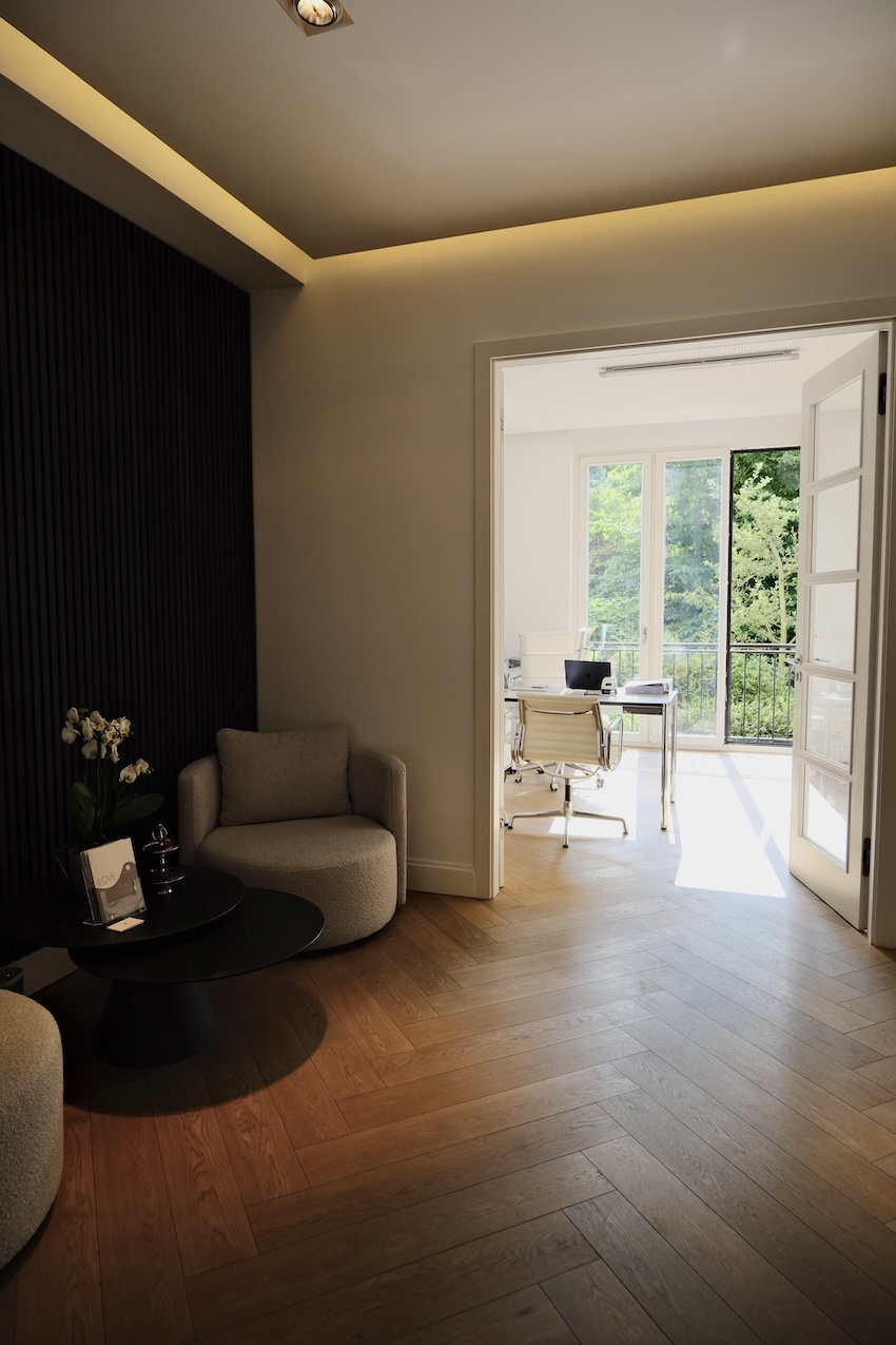 Moderner Wartebereich mit beigen Sesseln, schwarzem Tisch und Blick in ein helles Büro mit Glasfenstern und weißem Stuhl.