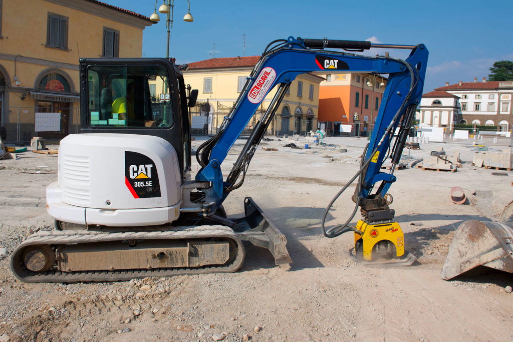 Mini escavatore CAT al lavoro per il rifacimento di una piazza cittadina con edifici storici sullo sfondo.