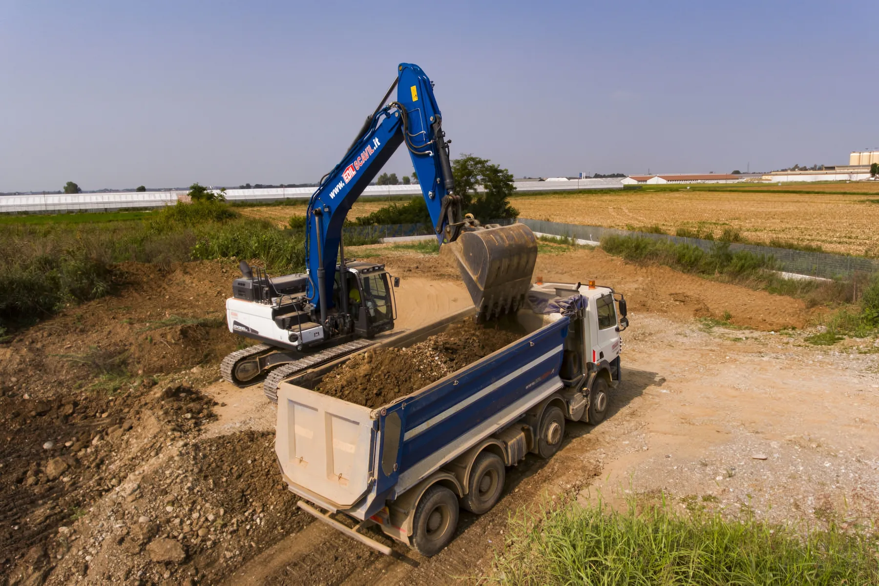 Escavatore cingolato blu che carica terra nel cassone di un autocarro in un ampio cantiere sterrato.