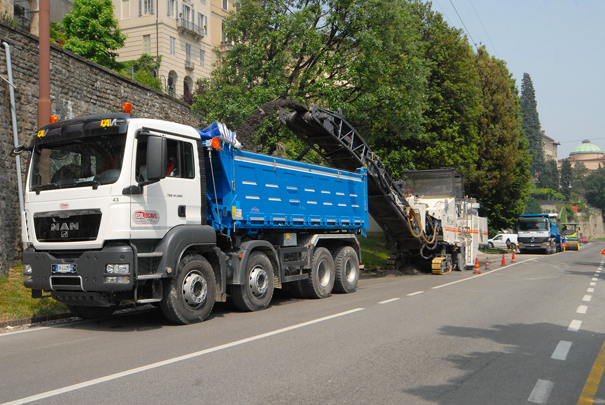 Autocarro MAN e fresatrice stradale in azione per il rifacimento del manto d'asfalto lungo una via alberata.