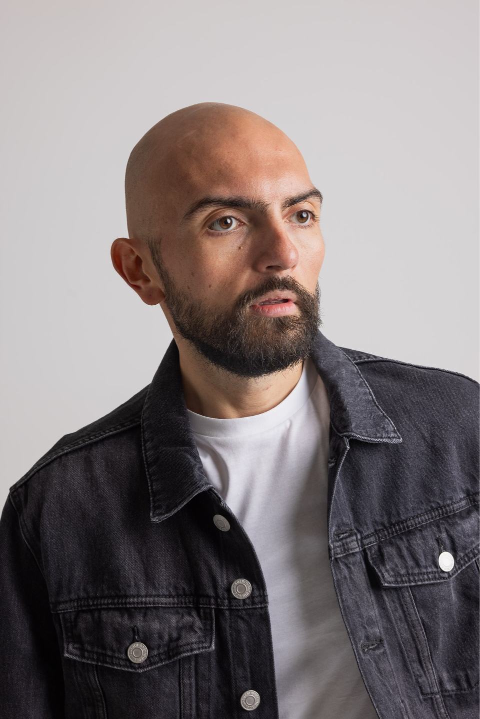 Portrait of a bald man with a beard wearing a black denim jacket and white t-shirt against a plain background.