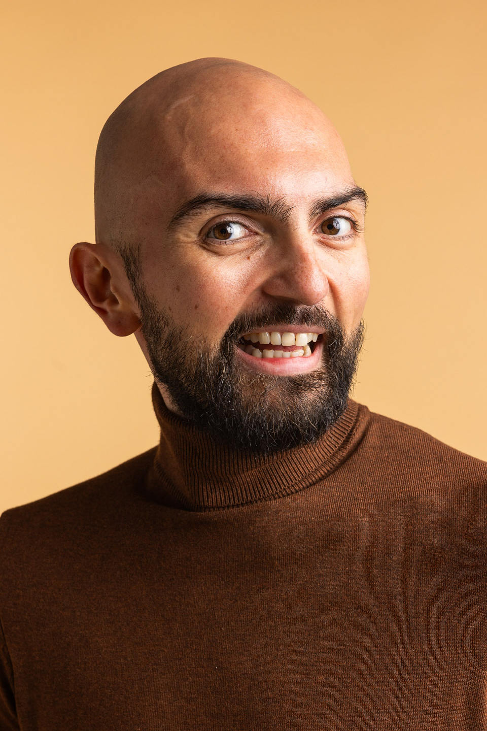Smiling bald man with a beard wearing a brown turtleneck sweater against a beige background.