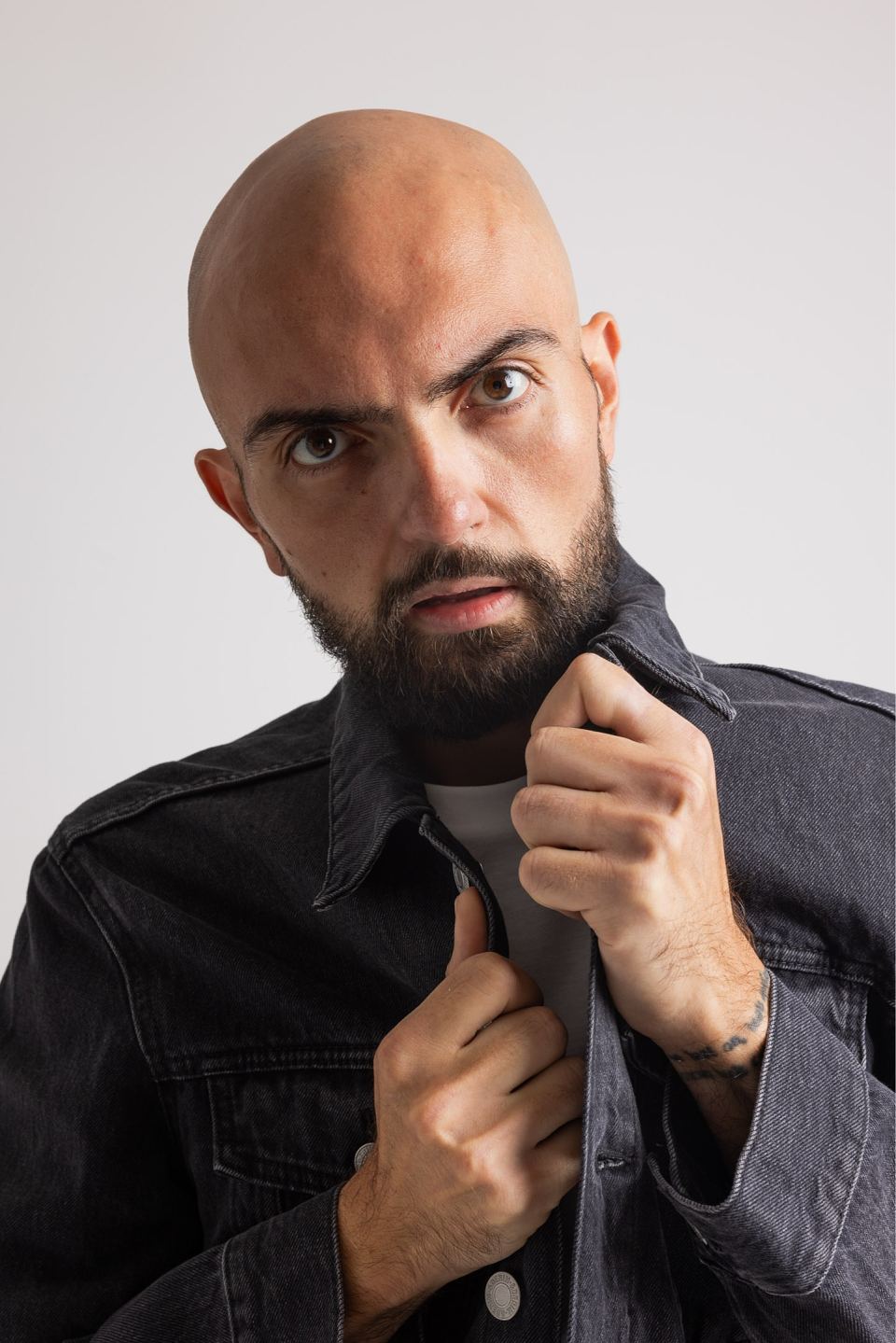 Serious bald man with a beard looking directly at the camera while adjusting the collar of his black denim jacket.