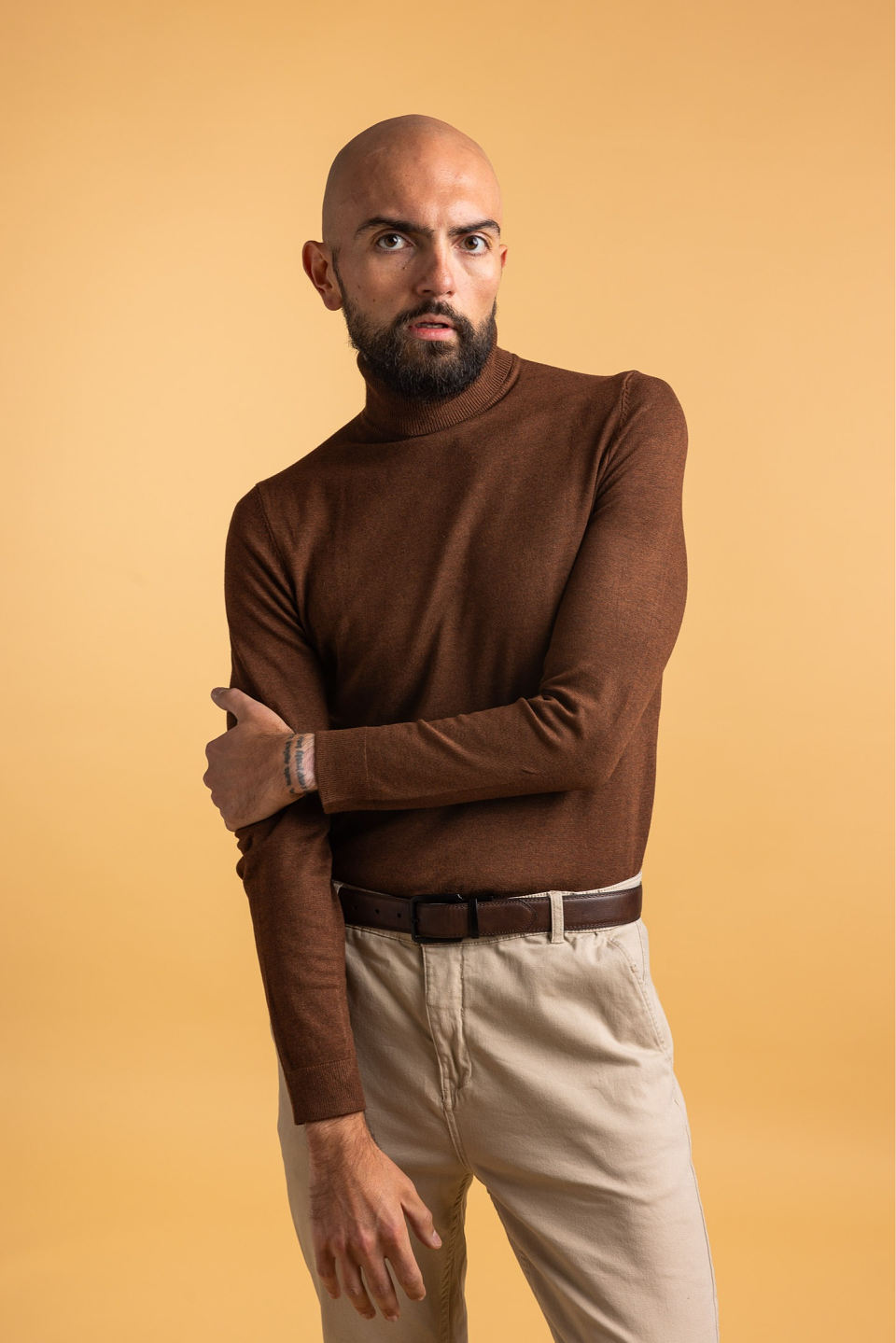 Bald man with beard wearing a brown turtleneck sweater and beige pants standing against a beige background.