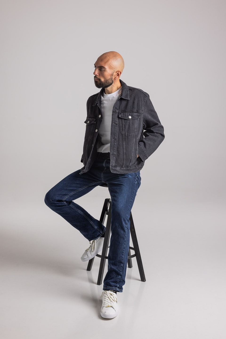 Bald man with beard wearing black denim jacket, white t-shirt, blue jeans, and white sneakers, sitting on a black stool against a plain background.