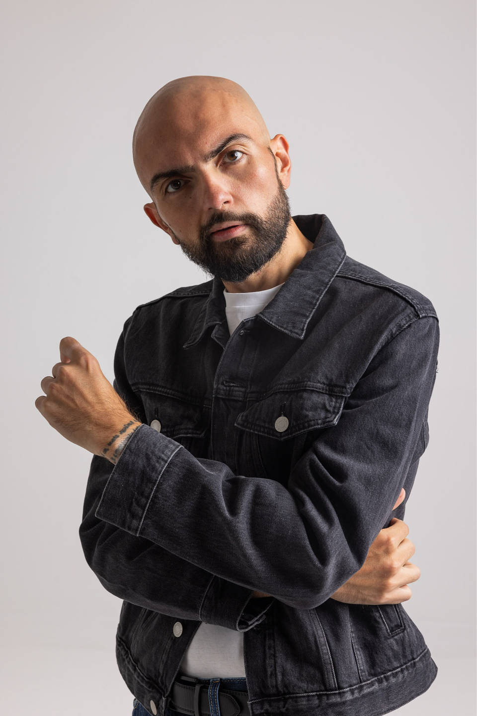 Bald man with beard wearing a black denim jacket and white shirt posing with crossed arms.