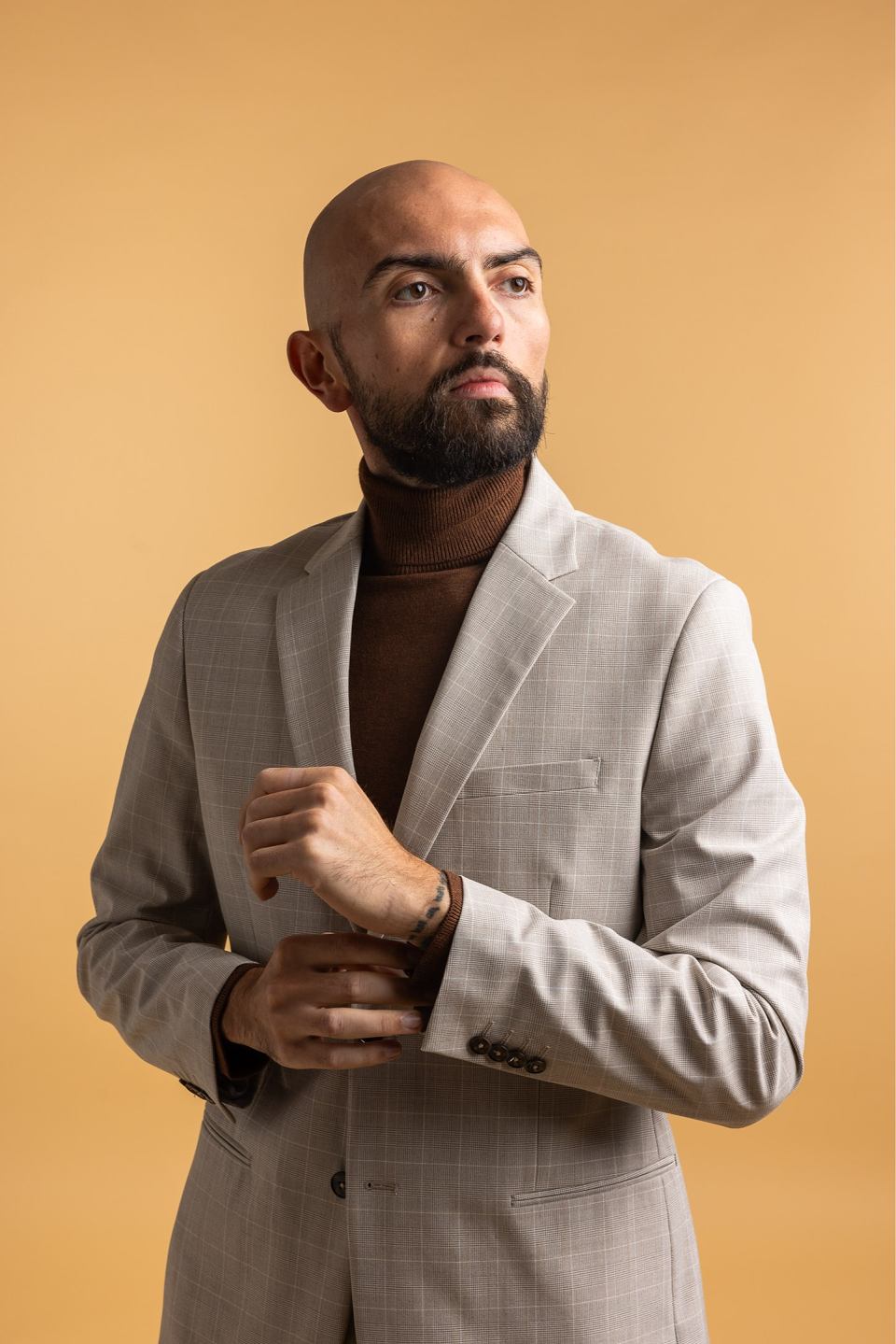 Bald man with a beard wearing a beige suit jacket and brown turtleneck, adjusting his sleeve against a plain warm beige background.