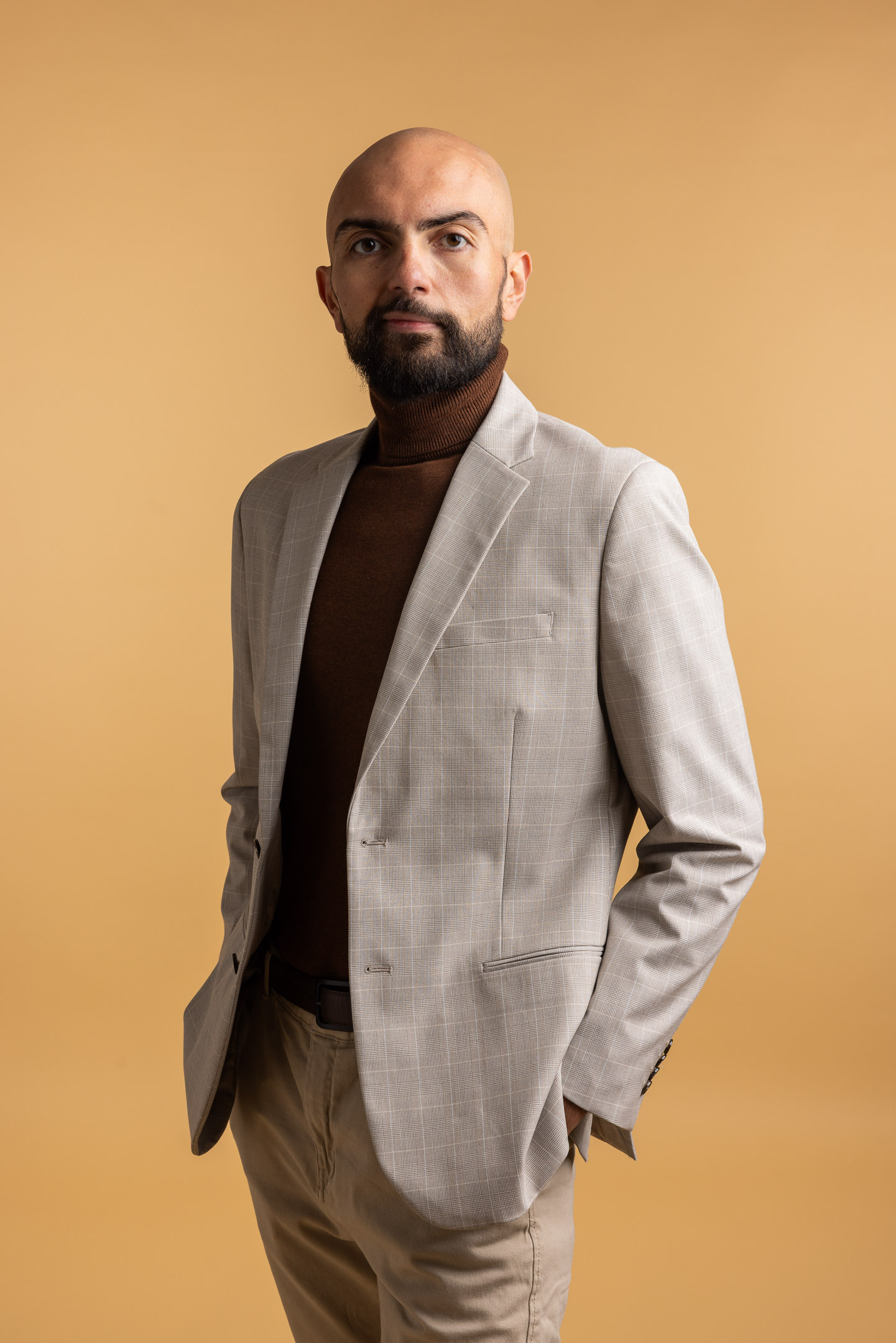 Serious bald man with beard wearing a light gray blazer, brown turtleneck, and beige pants against a light brown background.