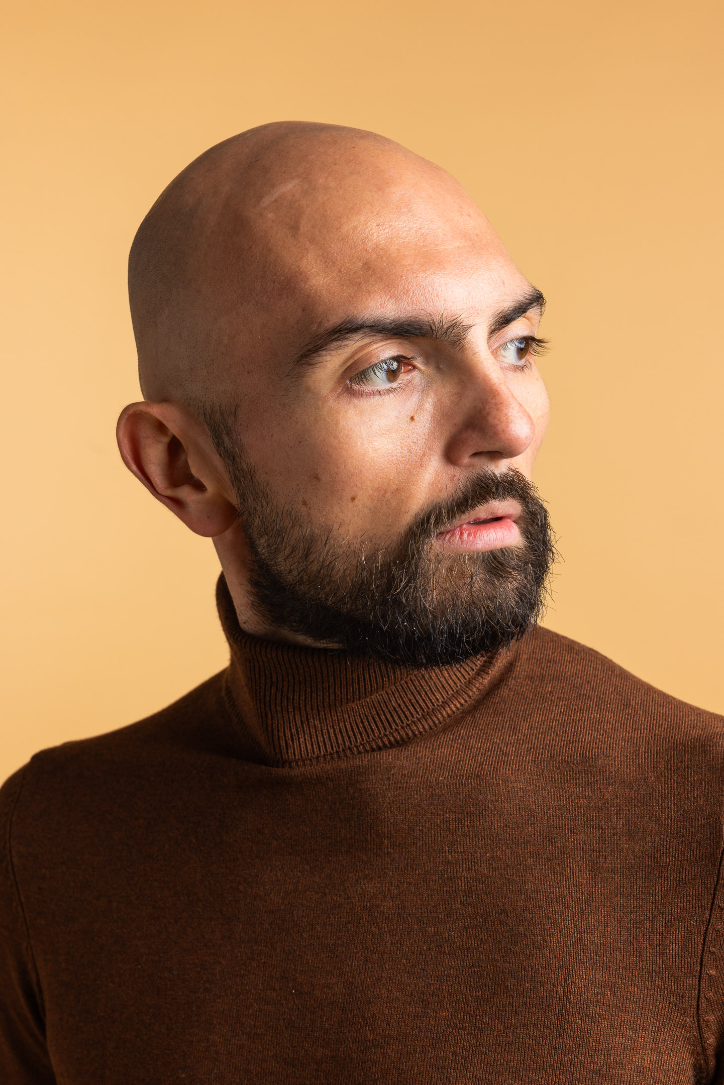 Close-up of a bald man with a beard wearing a brown turtleneck sweater against a beige background looking to the side.