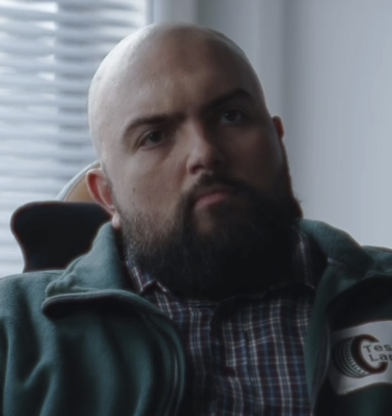 Portrait of a serious bald man with a beard wearing a green jacket and plaid shirt indoors.