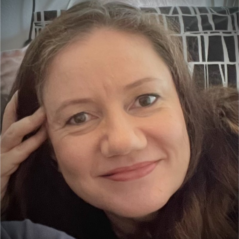 Close-up of a smiling woman with brown hair resting her head on her hand.