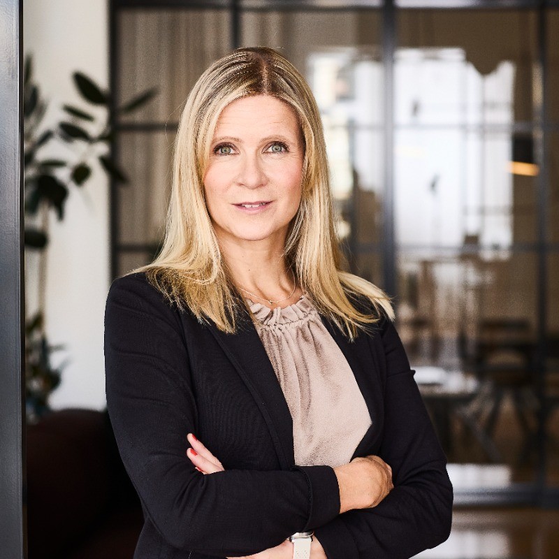 Professional woman with blonde hair wearing a black blazer and beige blouse standing with arms crossed in a modern office setting.