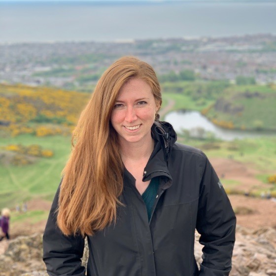 Smiling woman with long red hair wearing a black jacket standing outdoors with a scenic green landscape and a body of water in the background.