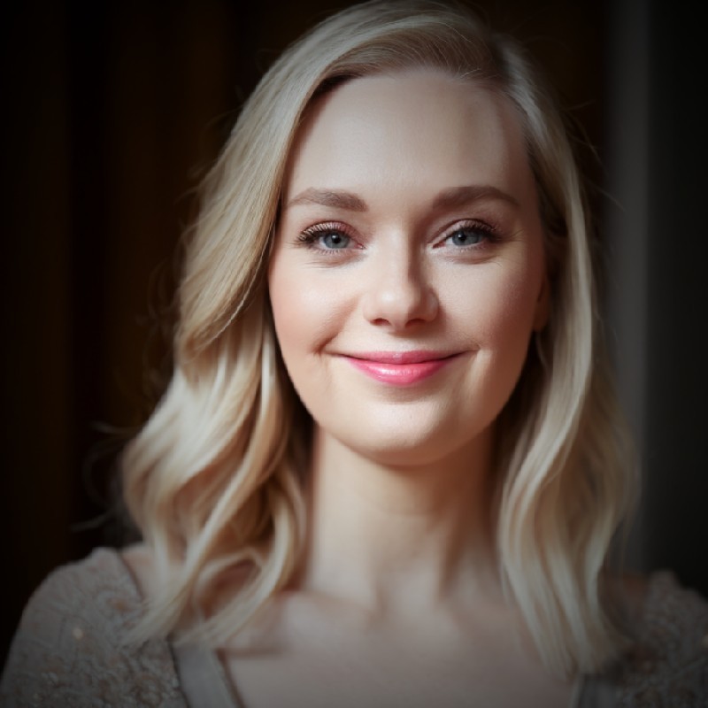 Smiling woman with shoulder-length blonde hair and light makeup against a dark blurred background.