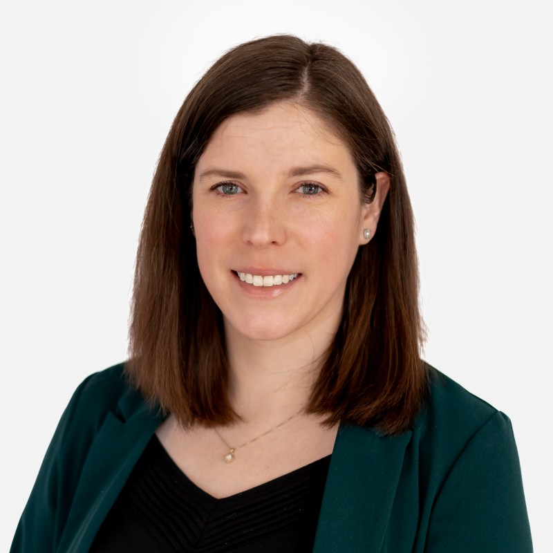Smiling woman with shoulder-length brown hair wearing a green blazer and black top against a white background.