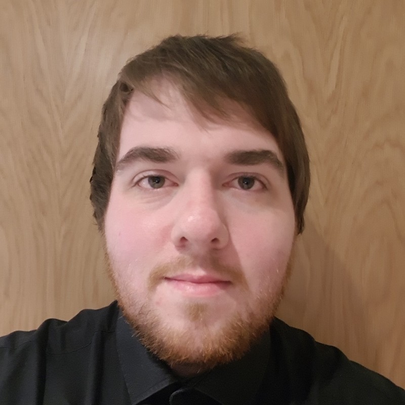 Headshot of a young man with short brown hair, light beard, and a black collared shirt in front of a wooden background.