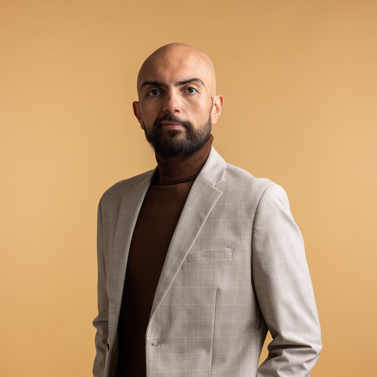 Bald man with a beard wearing a light gray plaid blazer and brown turtleneck standing against a beige background.