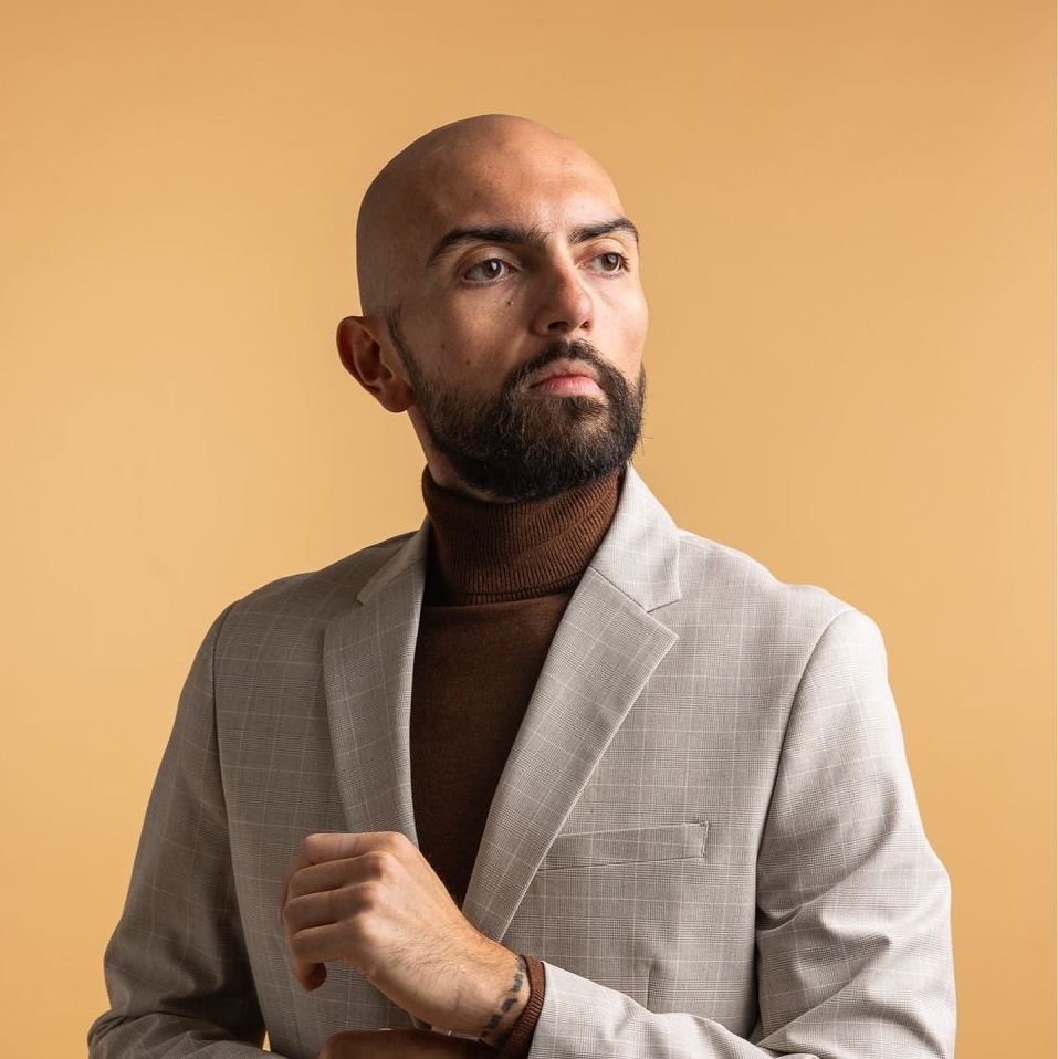 Bearded bald man in a beige blazer and brown turtleneck sweater adjusting his cuff against a beige background.