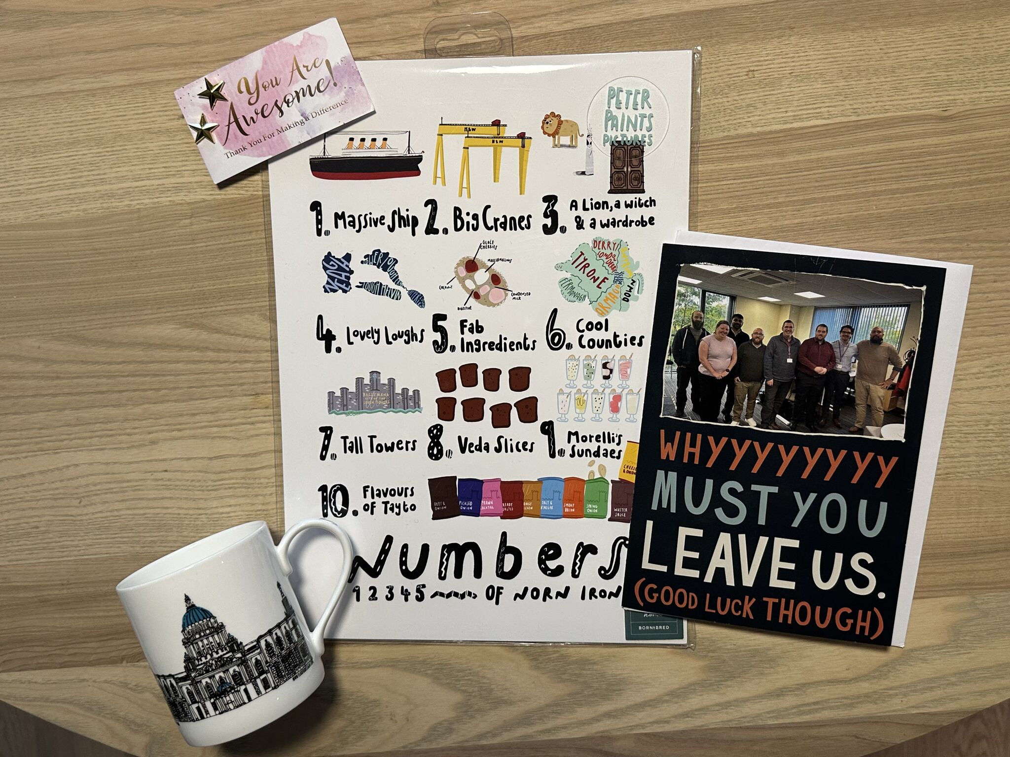 A white mug with a detailed black and blue illustration of a building, a colorful illustrated list titled 'Peter Paints Pictures' with ten labeled items, and a card with a group photo and text reading 'WHYYYYYYYY MUST YOU LEAVE US. (GOOD LUCK THOUGH)' on a wooden surface.