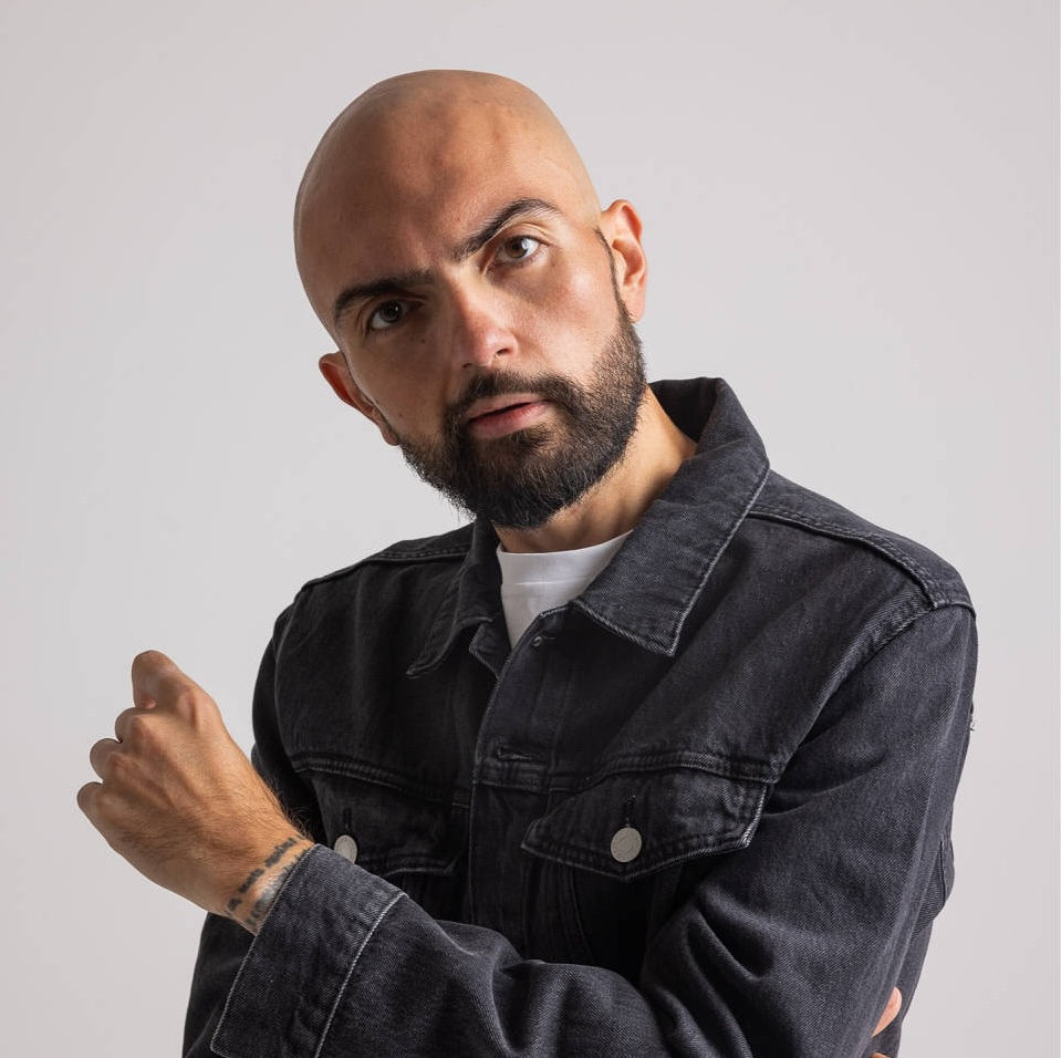 Bald man with a beard wearing a dark denim jacket and white shirt posing with his arm raised.