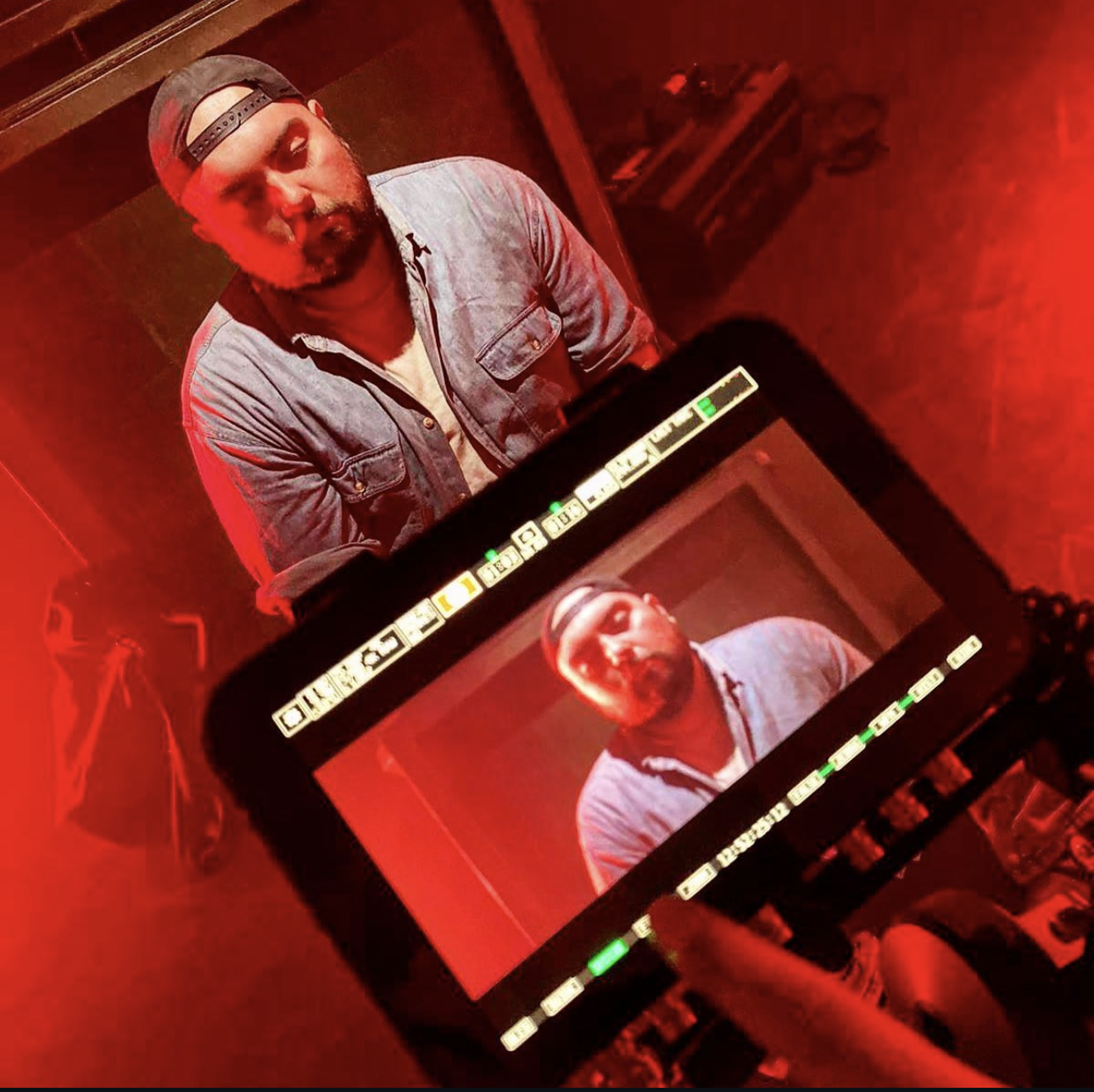 Bearded man in a backwards cap and denim shirt, seen on a camera monitor in a dimly lit red room.