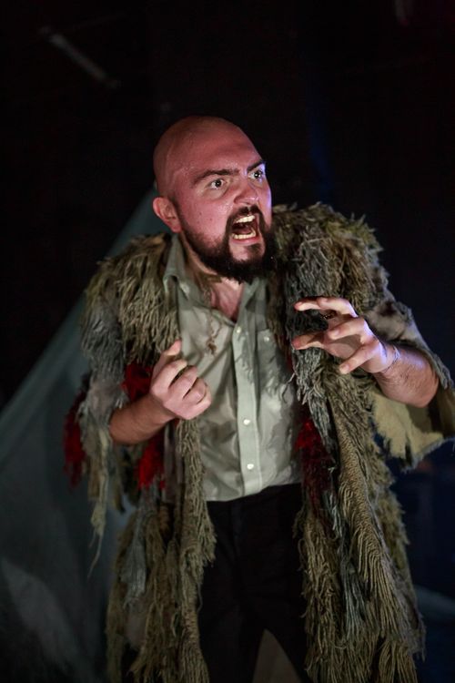 Angry man with a shaved head and beard wearing a worn, tattered cloak, gesturing fiercely in a dark setting.