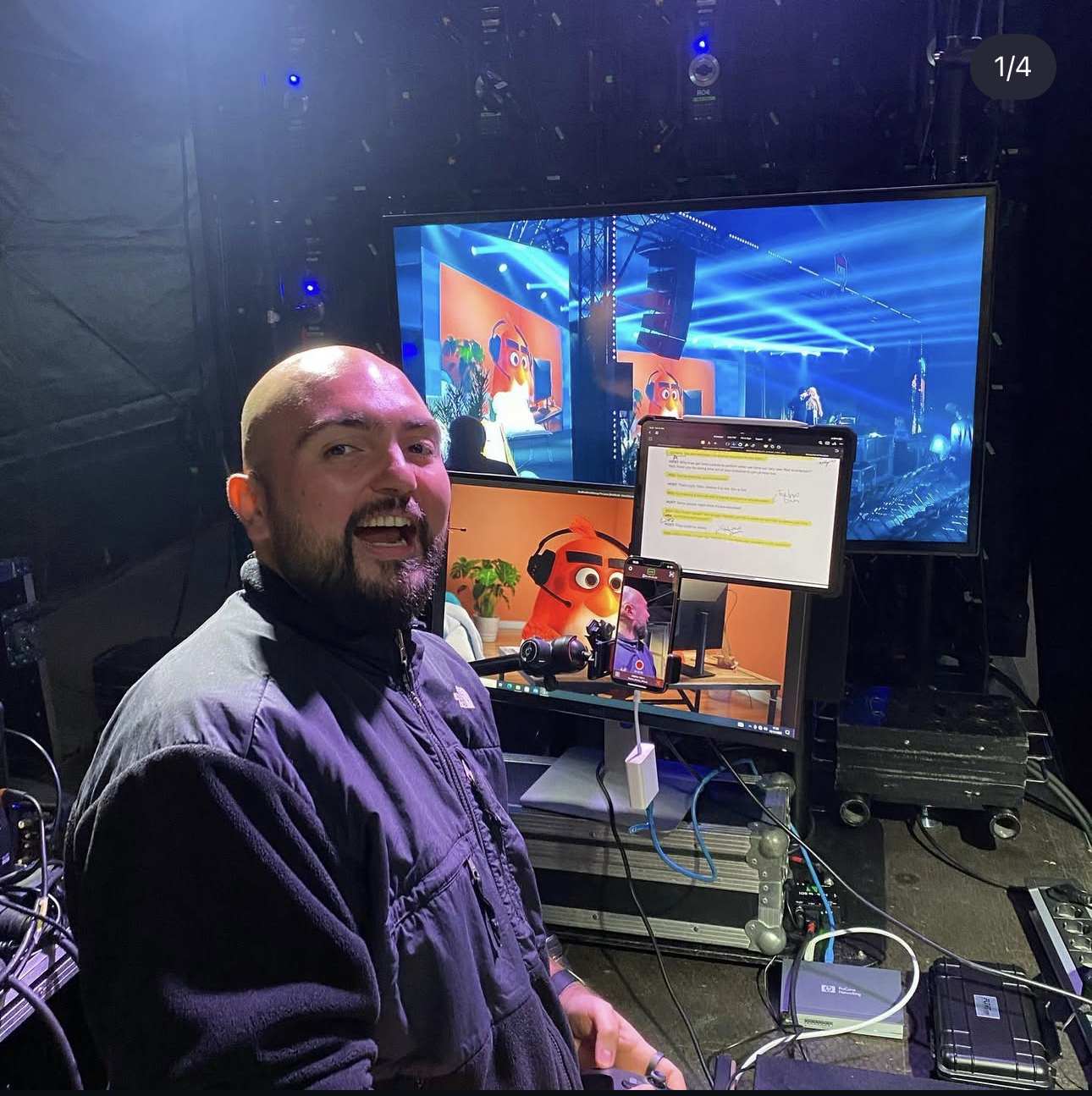 Smiling bald man in a black jacket sitting in front of multiple screens showing animated characters and a video call setup in a backstage tech area.