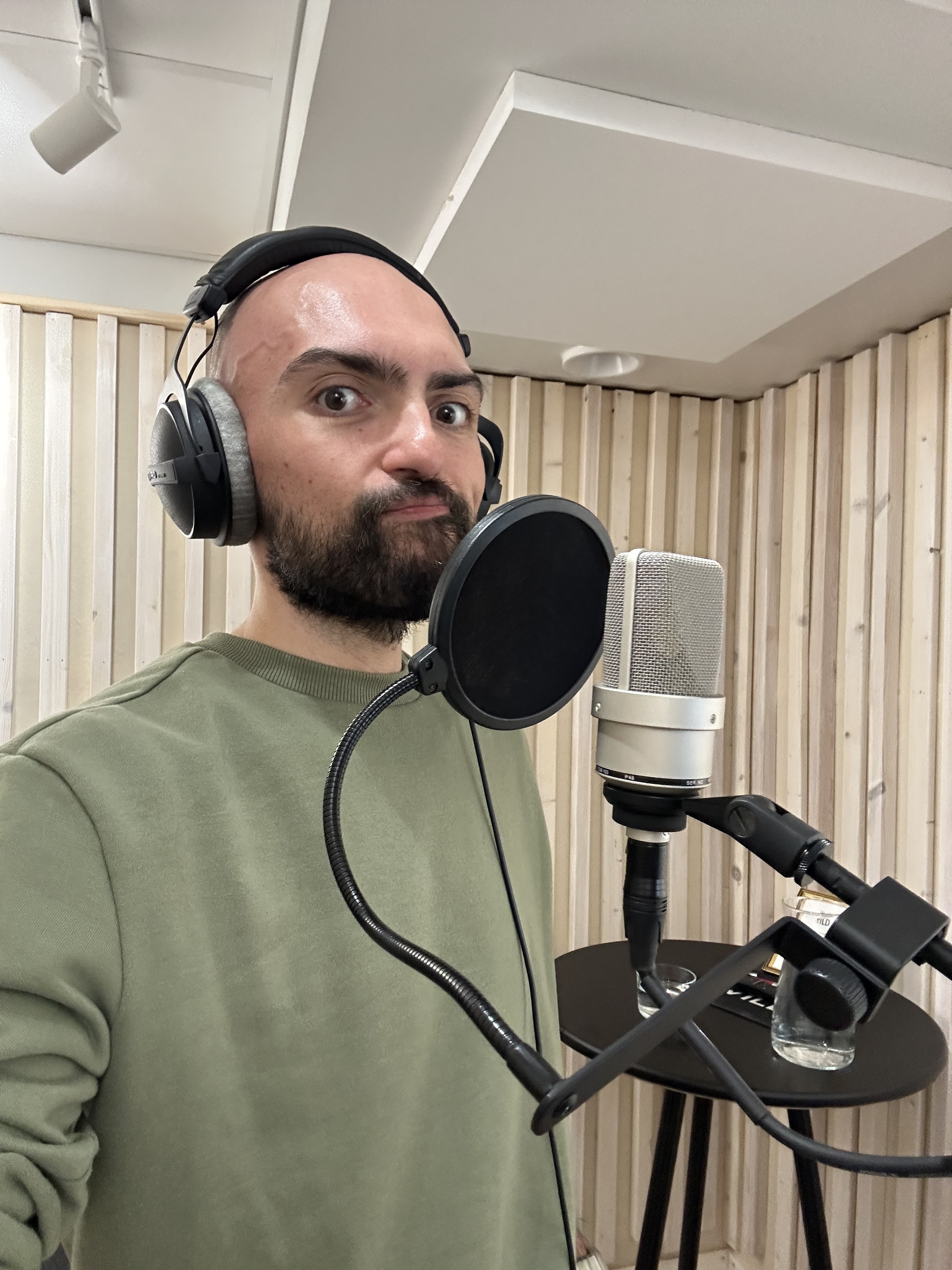 Man wearing headphones and a green sweatshirt standing in front of a microphone with a pop filter in a wooden-panelled room.